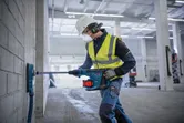 A person wearing safety equipment drills into a concrete wall with a rotary hammer.