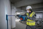 A person wearing safety equipment uses a rotary hammer to drill into a concrete wall.
