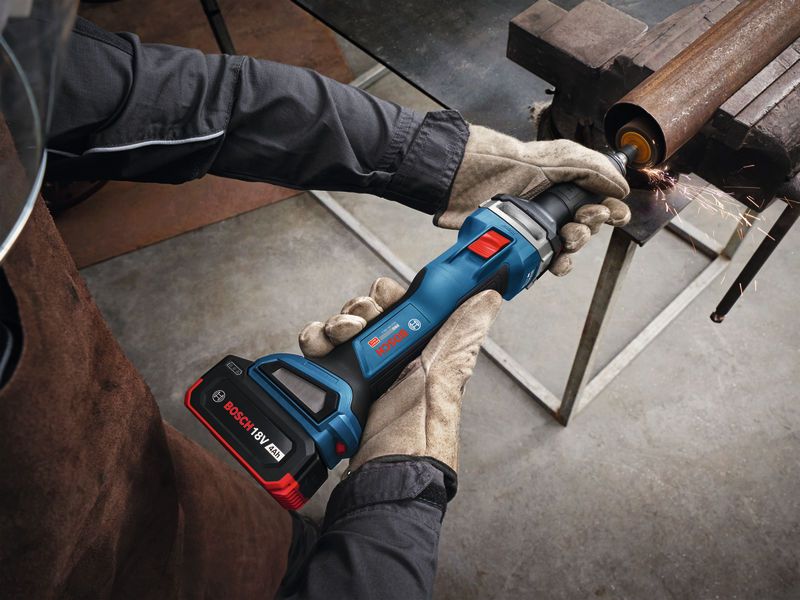 A person wearing safety equipment uses a cordless straight grinder to grind a metal pipe.