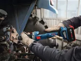 A person wearing safety equipment uses a cordless straight grinder on metal machinery.