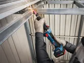 A person wearing safety equipment uses a cordless straight grinder on a metal beam.