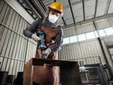 A person wearing safety equipment uses a cordless straight grinder on a metal beam.
