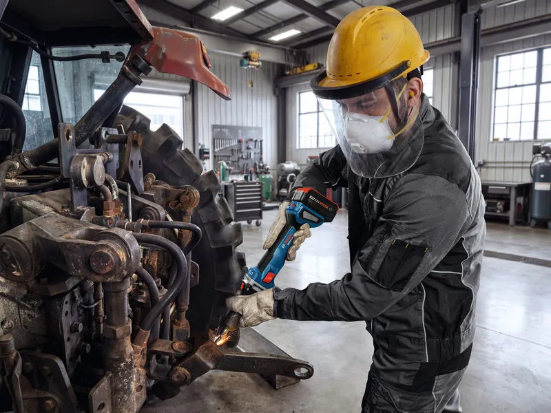 A worker wearing safety equipment uses a cordless grinder on heavy machinery.