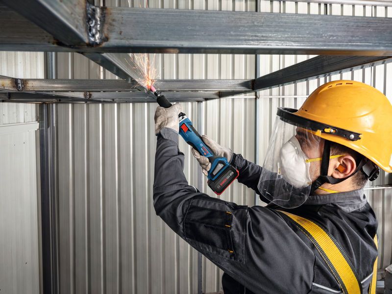 A person wearing safety equipment grinds a metal frame with a cordless straight grinder.