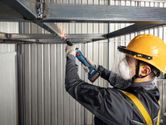 A person wearing safety equipment grinds a metal frame with a cordless straight grinder.