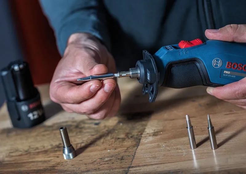 A person attaches a metal tip to a cordless soldering iron on a wooden surface.