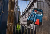 A blue and red power tool rests against a metal wire fence outdoors.