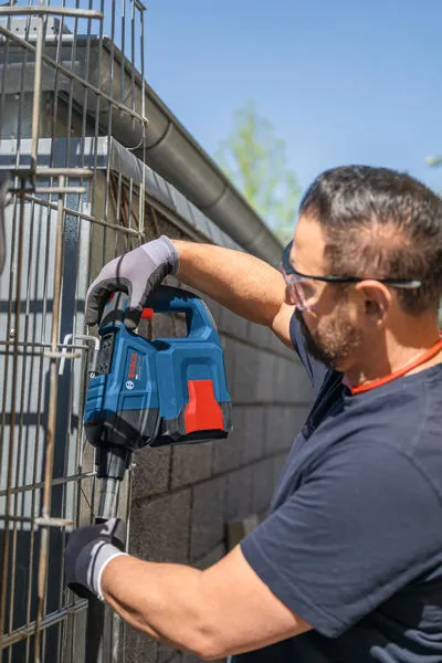 A person wearing safety equipment uses a power tool to drill into a metal fence.
