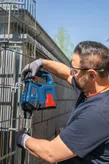 A person wearing safety equipment uses a power tool to drill into a metal fence.