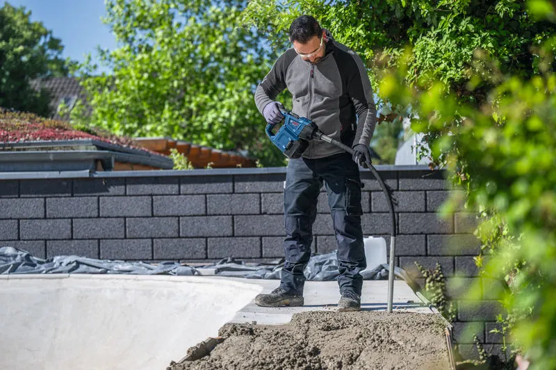 A person wearing safety equipment uses a power tool to compact concrete outdoors.