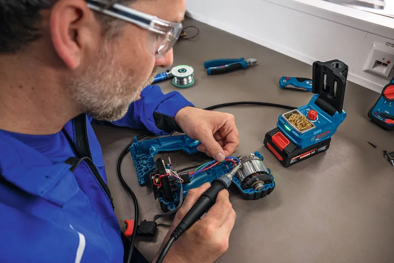 A person wearing safety equipment solders wires inside a power tool using a cordless soldering iron.
