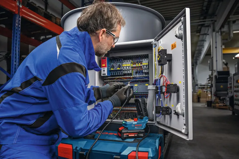A person wearing safety equipment solders wires inside an open electrical control panel.