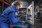 A person wearing safety equipment solders wires inside an open electrical control panel.