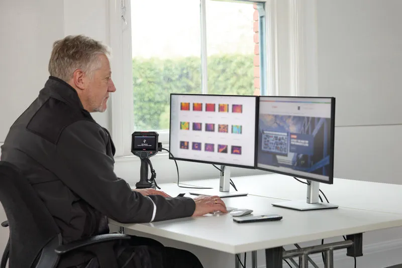 A person wearing safety equipment analyzes thermal images on dual monitors using a thermo camera.