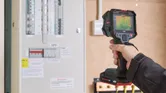 Person wearing safety equipment scans an electrical panel with a thermo camera.