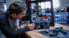 Person wearing safety equipment adjusts a 3D printer on a workbench with power tools nearby.