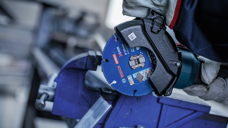 A person wearing safety equipment uses a power tool to cut metal clamped in a vise.