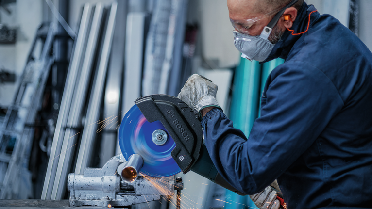 A person wearing safety equipment cuts a metal pipe with a circular saw, creating sparks.