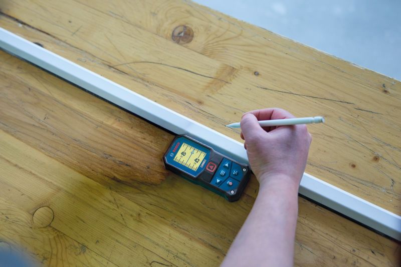 A person uses a laser measure to mark a line on a wooden surface.