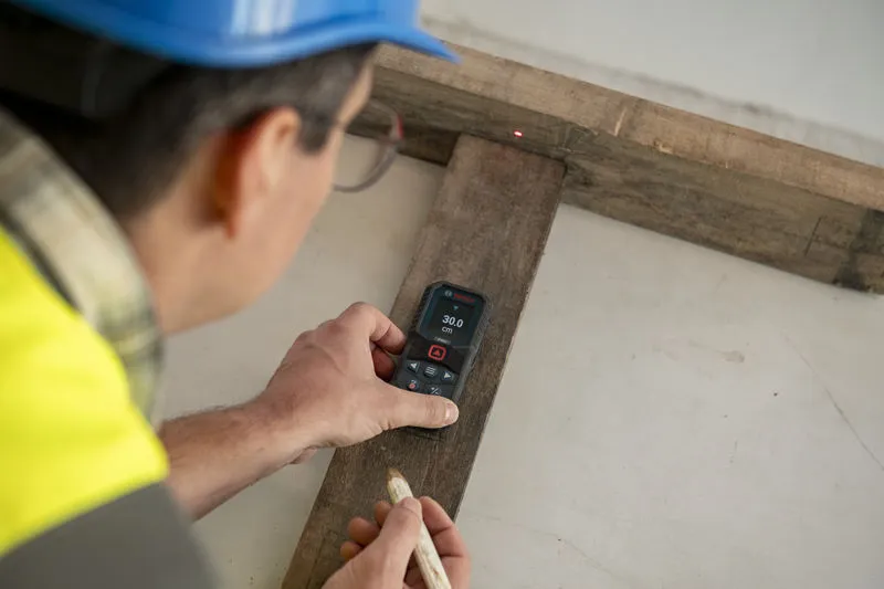 A person wearing safety equipment uses a laser measure to record wood length.