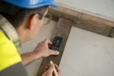 A person wearing safety equipment uses a laser measure to record wood length.