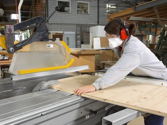 Worker wearing safety equipment cuts wood on a table saw in a carpentry workshop.