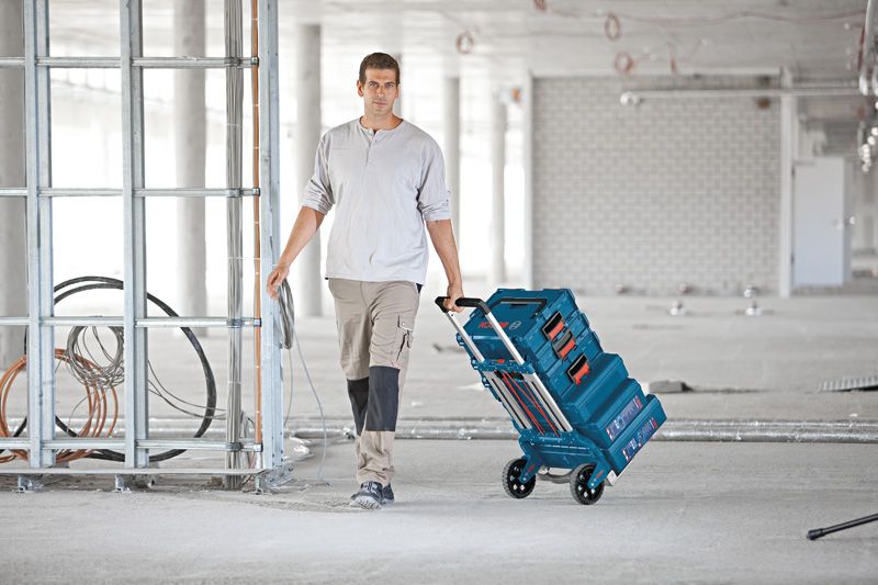 A person pulls a collapsible hand truck with stacked toolboxes in a construction area.