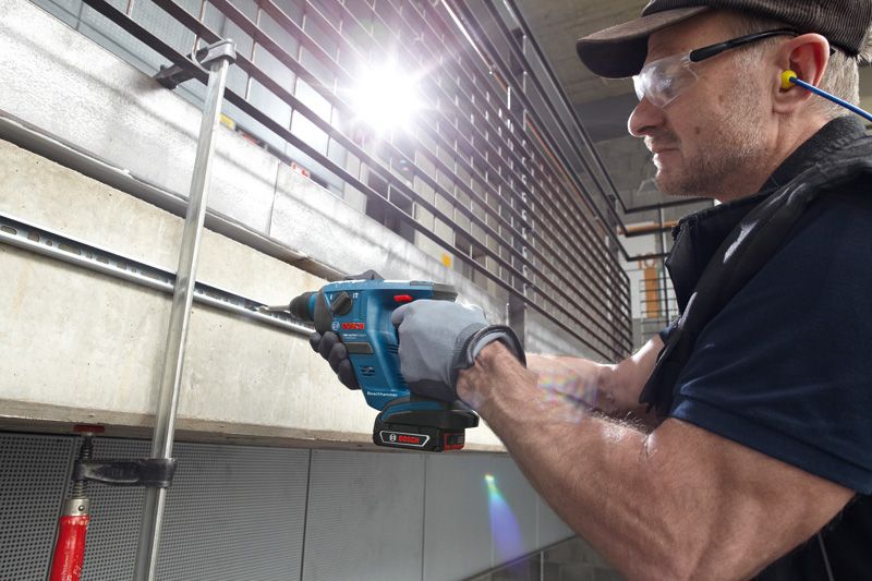 A person wearing safety equipment drills into concrete using a cordless rotary hammer.