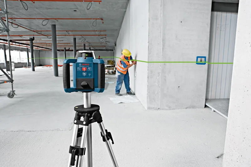A person wearing safety equipment marks a wall using a laser leveling tool on a tripod.