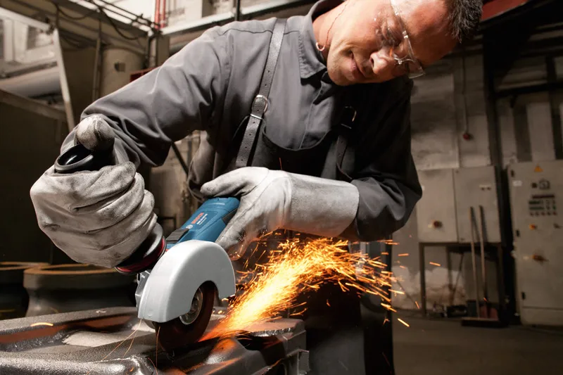 A person wearing safety equipment grinds metal with an angle grinder, producing sparks.