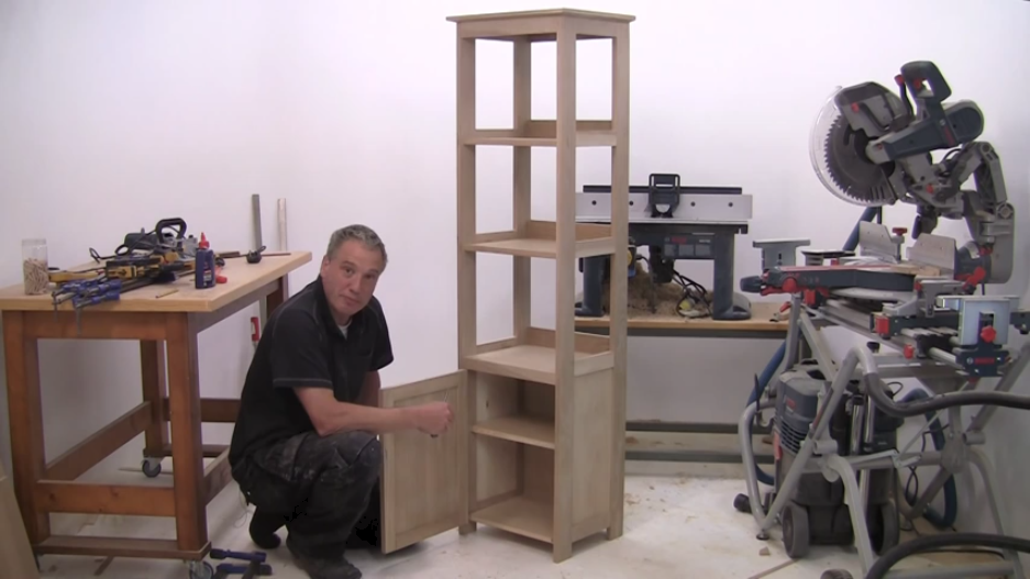 A man kneels beside a wooden shelf in a workshop with tools and machines.