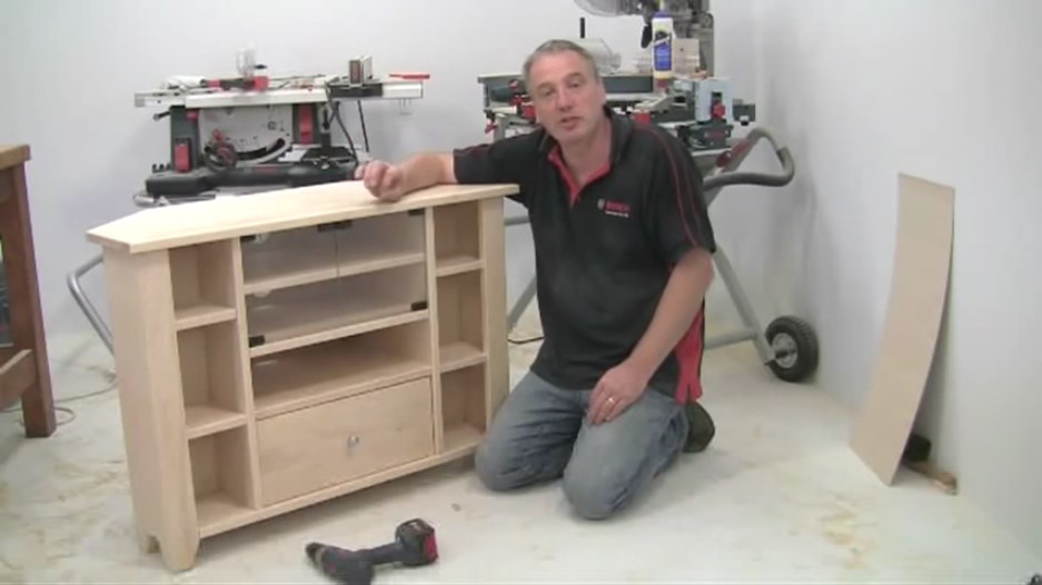 A man in a black Bosch shirt kneels by a wooden TV stand in a workshop.