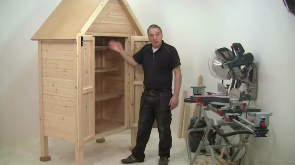 A man gestures beside a wooden shed and a miter saw in a workshop setting.