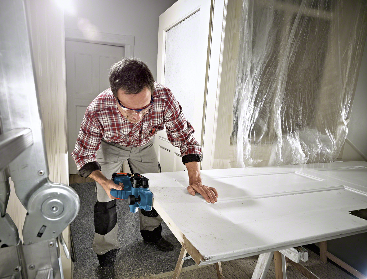 A man in glasses uses a blue Bosch tool on a white door in a bright room.