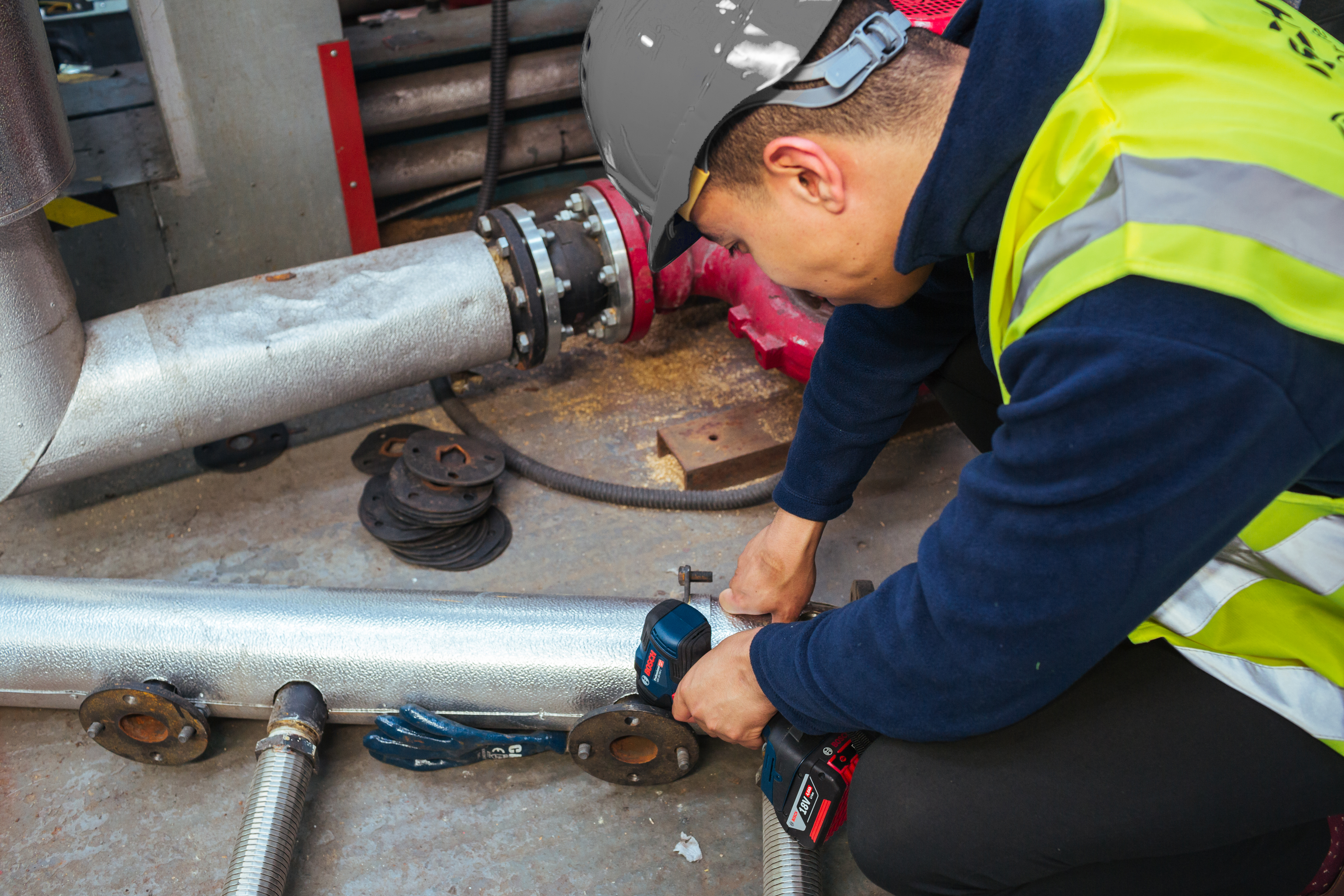 A worker in a helmet tightens a pipe with a tool in a workshop setting.