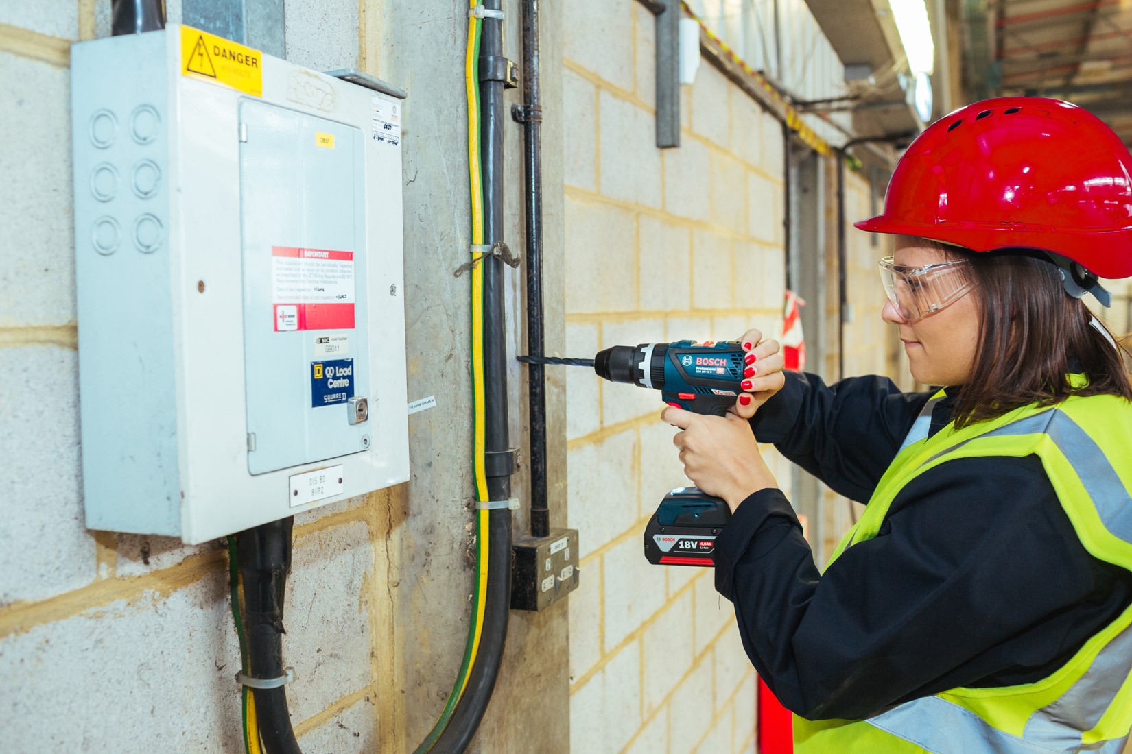 A woman in a hard hat uses a Bosch Professional drill on a wall near an electrical box.