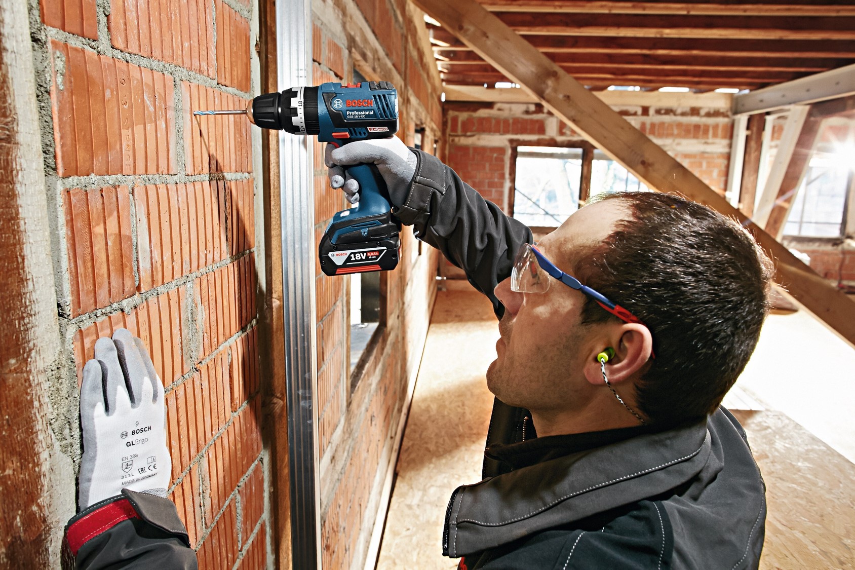 A man drills into a brick wall with a Bosch Professional drill, wearing gloves and glasses.