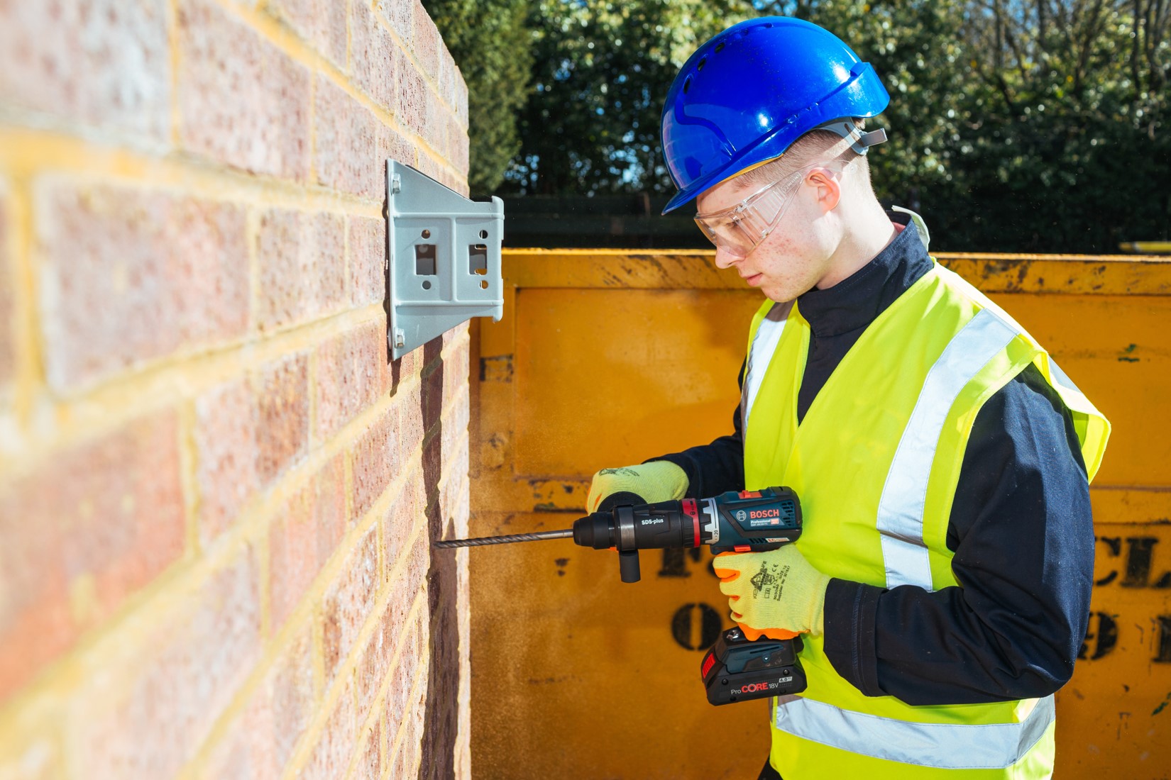 A worker in a yellow safety vest drills into a brick wall with a Bosch drill.