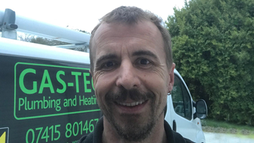 A smiling man stands in front of a white van with a green logo and trees behind him.
