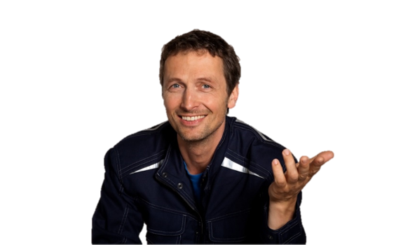 A smiling man in a blue jacket gestures with his hand, against a plain background.
