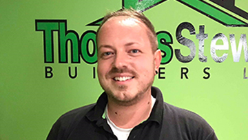 A smiling man in a black polo shirt stands against a green wall with logos.