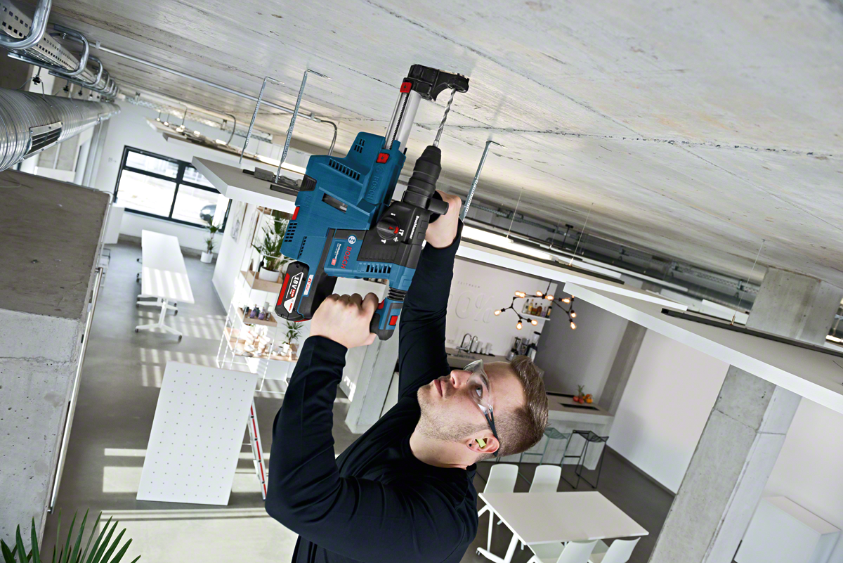A man uses a blue Bosch Professional rotary hammer drill on a ceiling in a modern workspace.