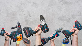 Hands hold various Bosch Professional power tools against a gray wall background.
