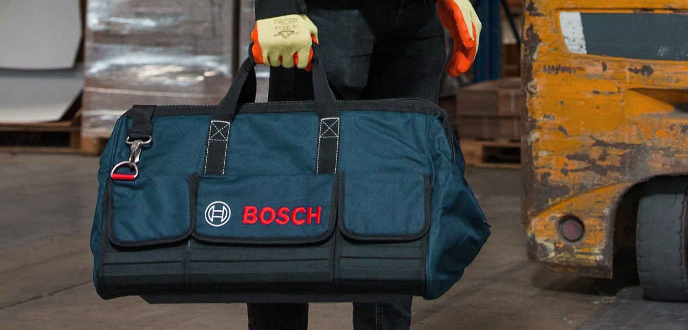 A person carries a dark blue Bosch tool bag in a warehouse setting.