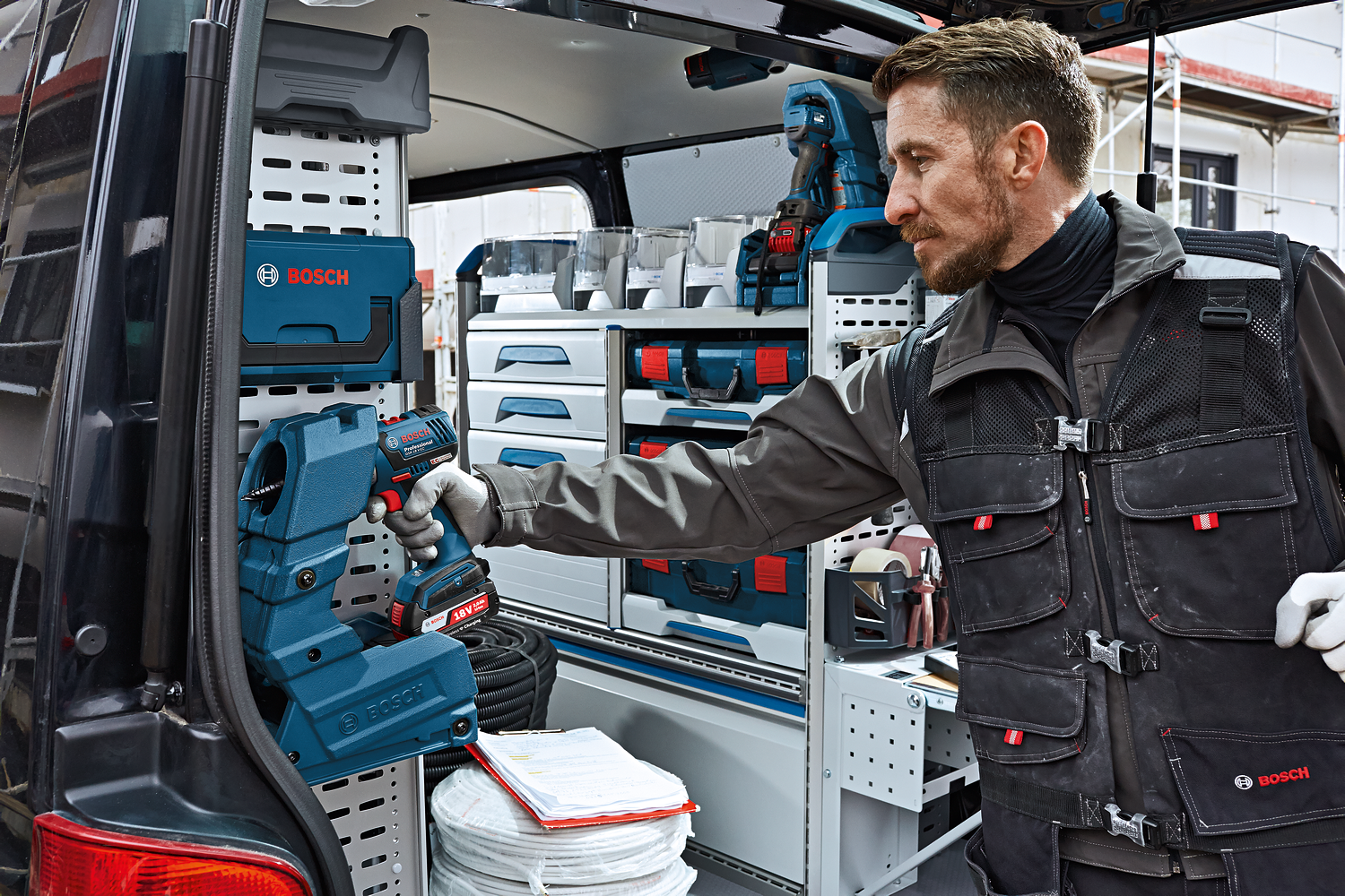 A man in gloves holds a Bosch Professional drill in a van filled with tools and storage boxes.