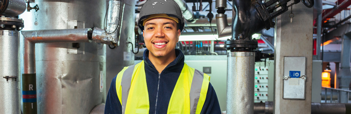A smiling worker in a helmet and yellow vest stands in a factory with pipes around.