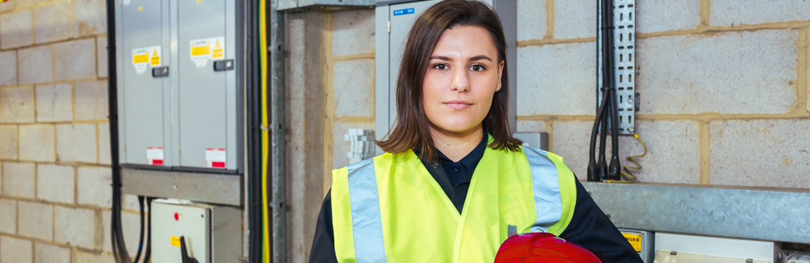 A woman in a yellow safety vest stands in a workshop with electrical panels behind her.