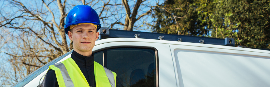 A young man in a blue hard hat and reflective vest stands by a white van outdoors.