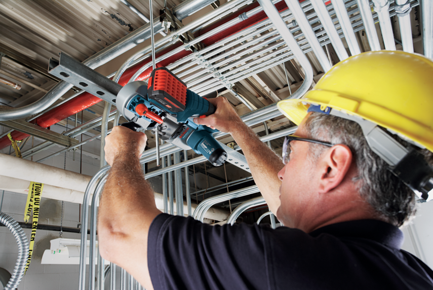 A worker in a yellow hard hat uses a Bosch Professional tool on pipes above him.