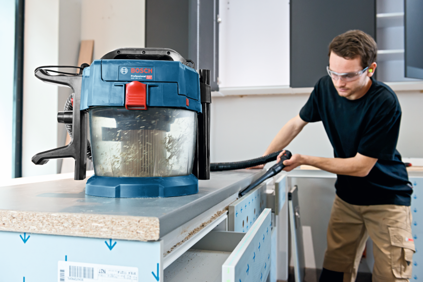A blue Bosch Professional vacuum is used by a man in a black shirt in a kitchen.